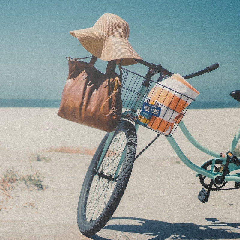 The Rowdy Leather Crossbody Bag hanging from the handlebars of a bicycle on the beach with a straw hat