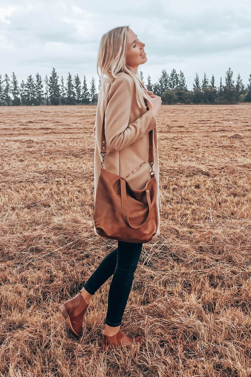 Woman in a camel coat and ankle boots wearing the Rowdy Leather Crossbody Bag across the body in a field