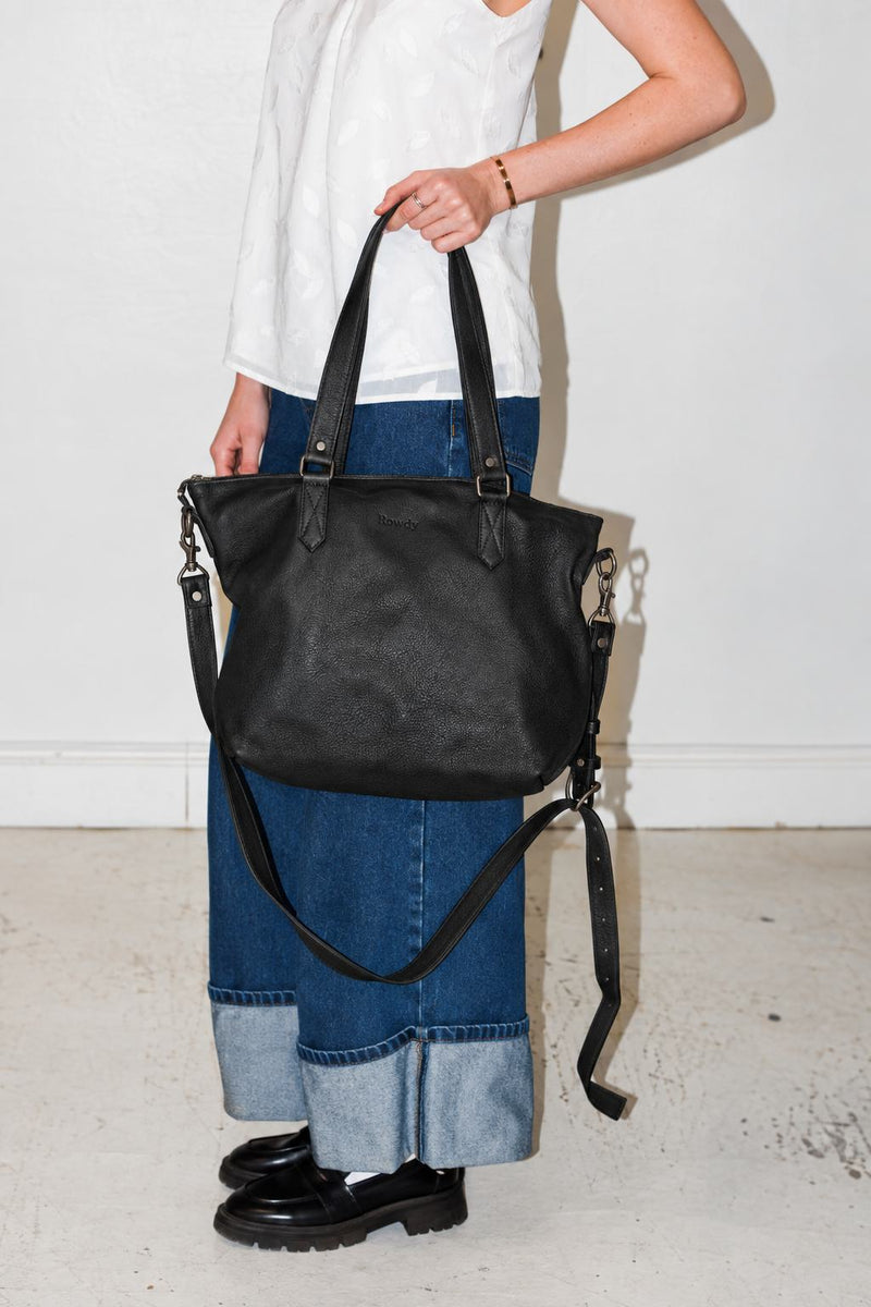 Woman in a white top and wide cuffed jeans holding the Rowdy Leather Crossbody Bag in Charcoal by its handles