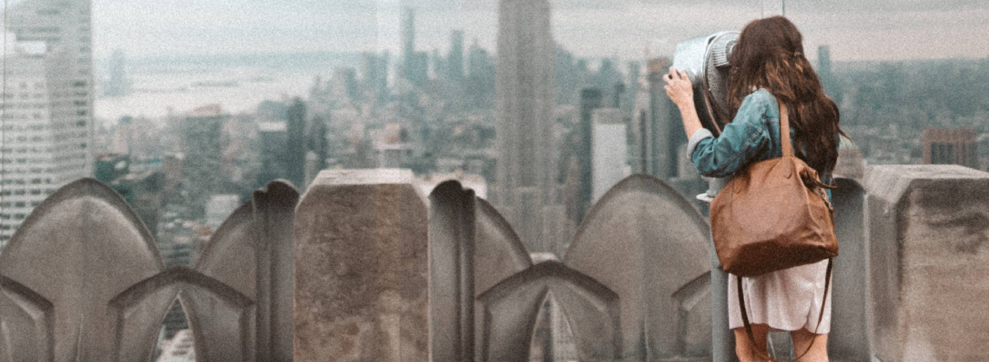 Woman in a denim jacket looking out over a city skyline through rooftop binoculars, with the Rowdy Leather Crossbody Bag on her shoulder