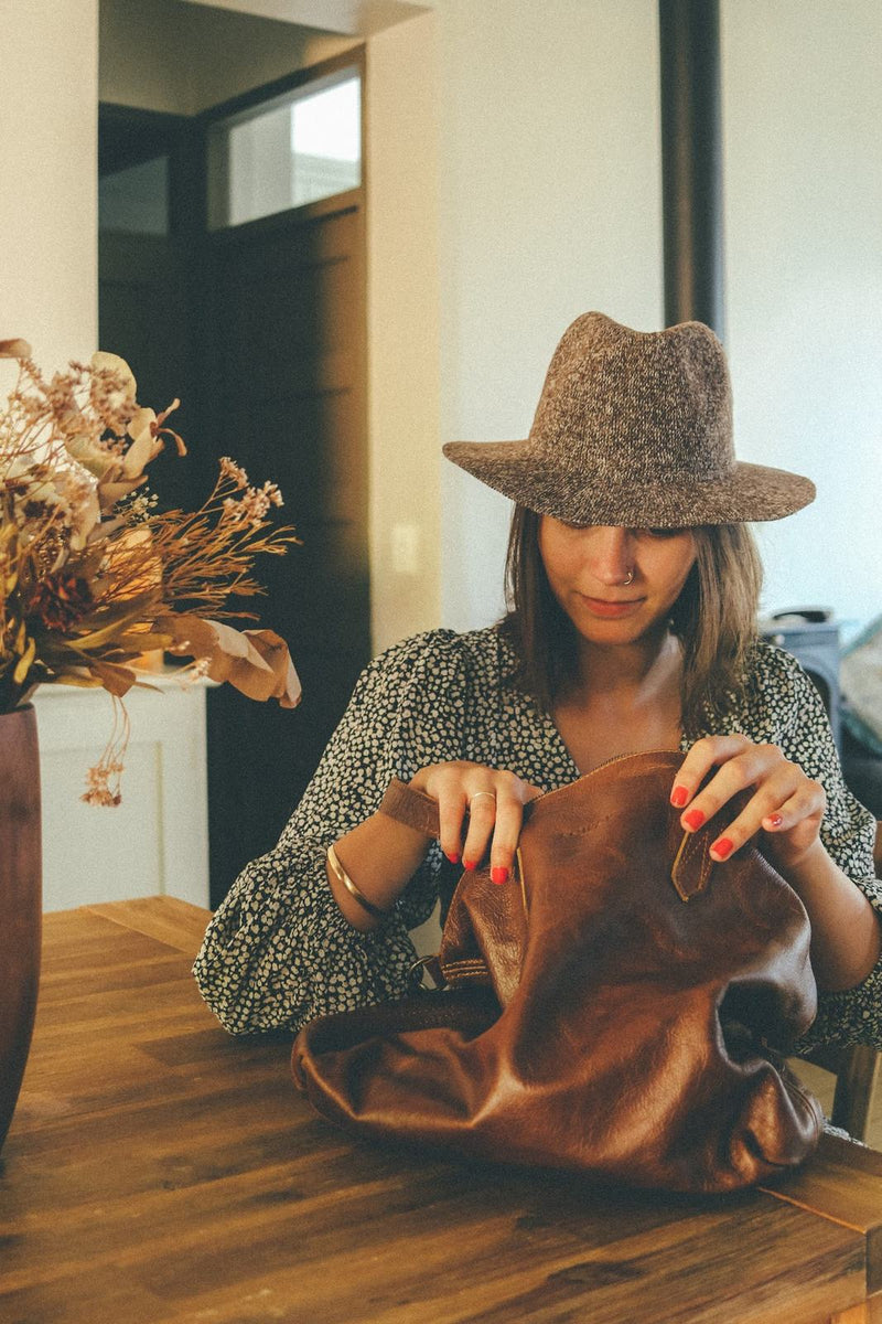 Woman in a felt hat and patterned blouse opening the Rowdy Leather Crossbody Bag on a café table