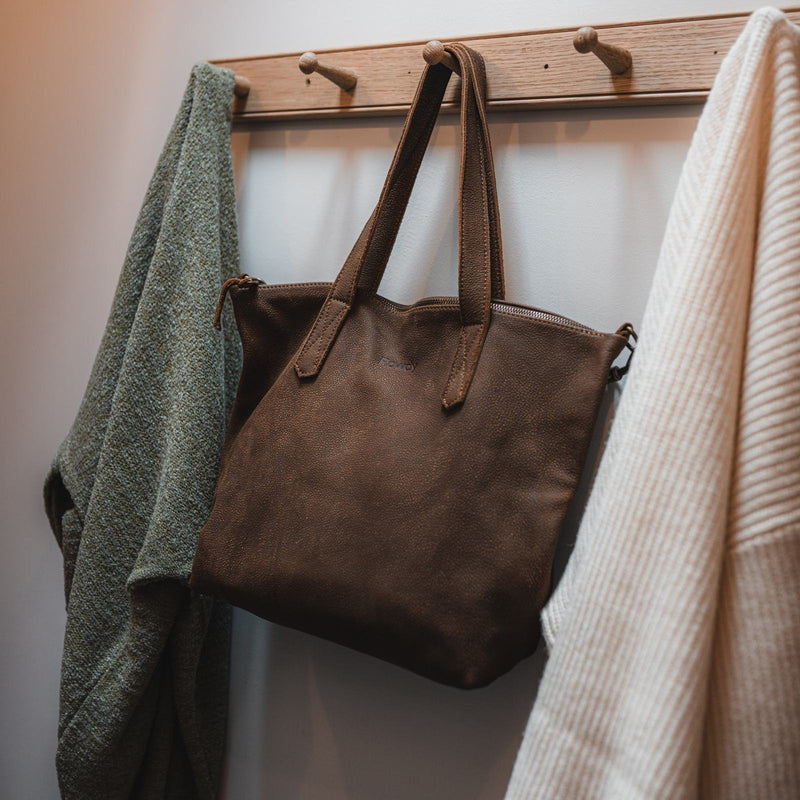 The Rowdy Leather Crossbody Bag hanging on a peg rail between a green and a cream knit