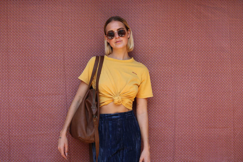 Woman in a knotted yellow T-shirt and navy velvet trousers wearing the Rowdy Leather Crossbody Bag on her shoulder against a patterned wall
