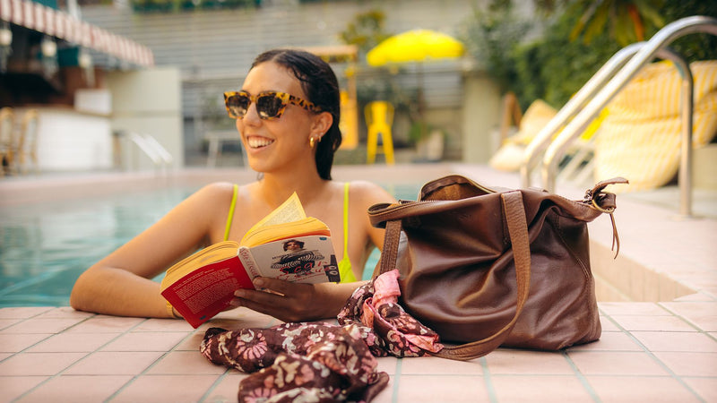 Woman reading in a pool, with a brown Rowdy Leather Tote Bag and a silk scarf on the poolside tiles