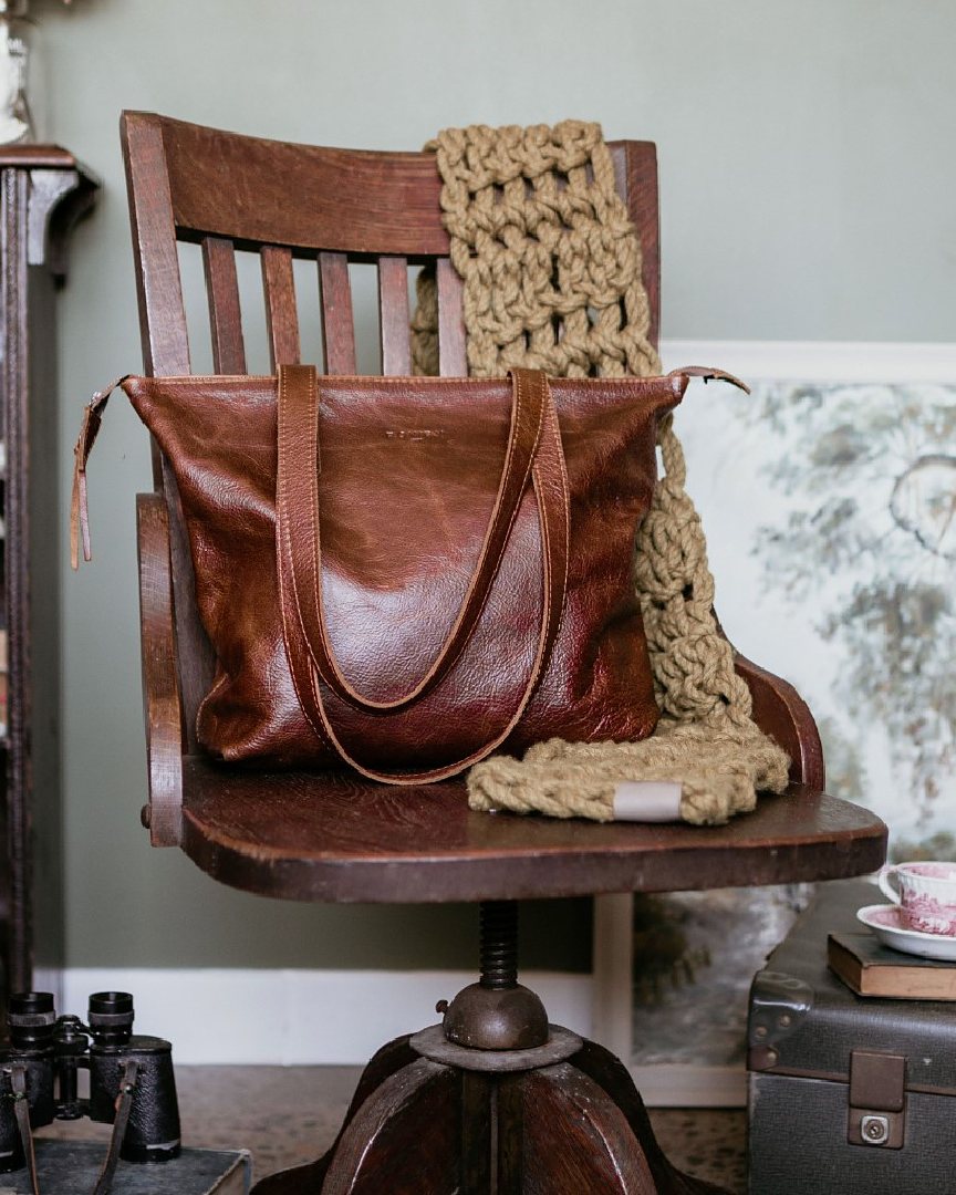 Rowdy Leather Tote Bag resting on a vintage wooden chair beside a chunky knit scarf, showing its long straps, zip top and embossed Rowdy mark